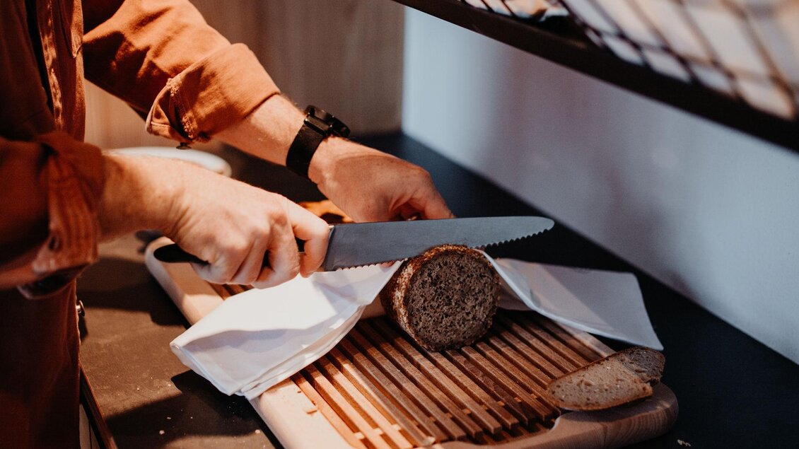 Eine Person schneidet frisches Brot auf einem Holzbrett. Im Hintergrund sind Körbe mit weiteren Broten sichtbar.