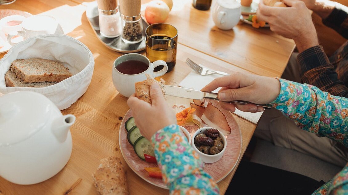 Ein gedeckter Frühstückstisch mit frischem Brot, Gemüse und Getränken. Eine Person schneidet gerade ein Brötchen auf.