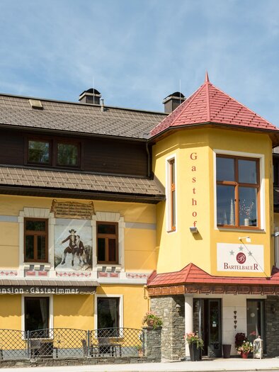 A charming inn with a yellow facade and a red roof. In front of the building are trees and an inviting terrace. | © Fred Lindmoser