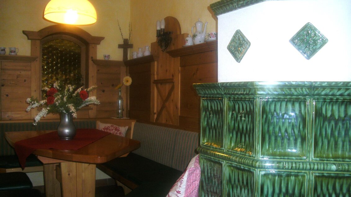 Ein einladender Raum mit Holzinterieur und einer gemütlichen Sitzecke. Im Mittelpunkt steht ein traditioneller Kachelofen in Grün. | © Gasthof Bartelbauer
