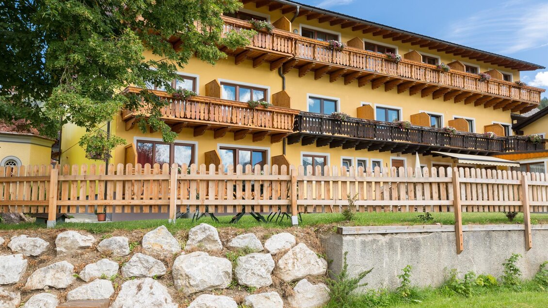 Ein mehrstöckiges, gelbes Gebäude mit Balkonen und Holzzaun. Im Vordergrund sind Steine und Gras zu sehen. | © Gasthof Bartelbauer / Fred Lindmoser