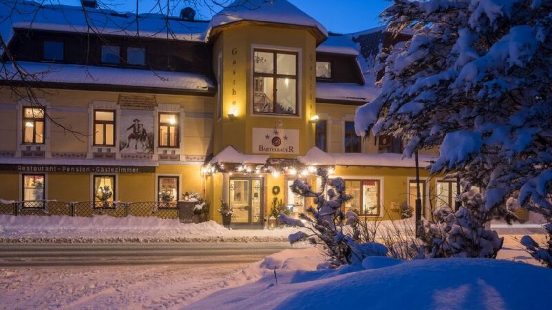 Ein gemütliches Hotel im Schnee bei Dämmerung. Die Fassade leuchtet warm und einladend. | © Gasthof Bartelbauer / Fred Lindmoser