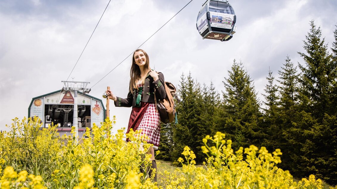 Eine junge Frau steht in einem gelben Blumenfeld. Im Hintergrund ist eine Seilbahn zu sehen, die durch einen bewaldeten Bereich fährt. | © Mariazeller Bürgeralpe / Fred Lindmoser