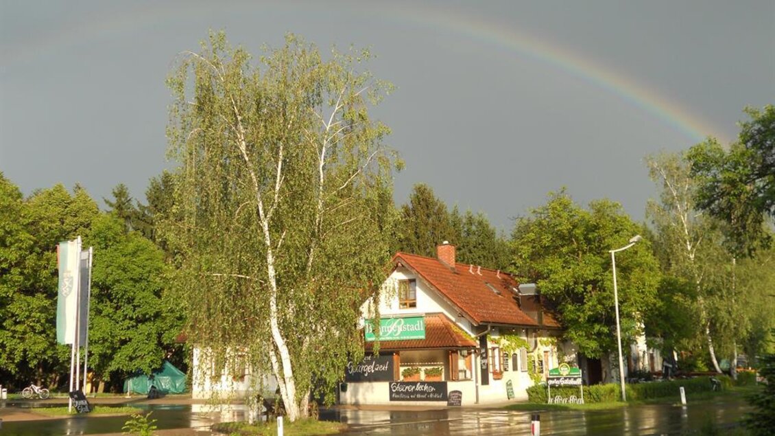 Ein ruhiges Straßenbild mit einem kleinen Gebäude und grünen Bäumen. Ein Regenbogen spannt sich über den Himmel nach einem Regenschauer.