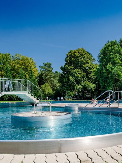 Ein schön gestalteter Außenpool mit klarem Wasser und einer kleinen Brücke. Umgeben von grünen Bäumen und einem blauen Himmel. | © Mias Photoart