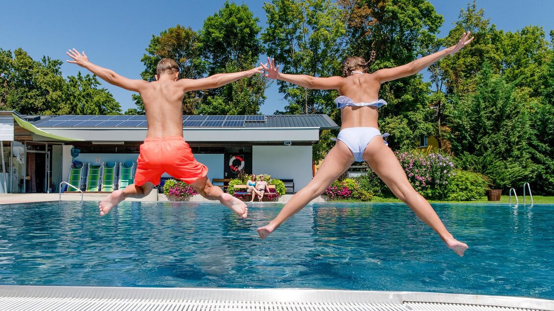 Zwei Personen springen gleichzeitig in einen klaren Pool. Die Sonne scheint und die Umgebung ist grün und einladend. | © Mias Photoart