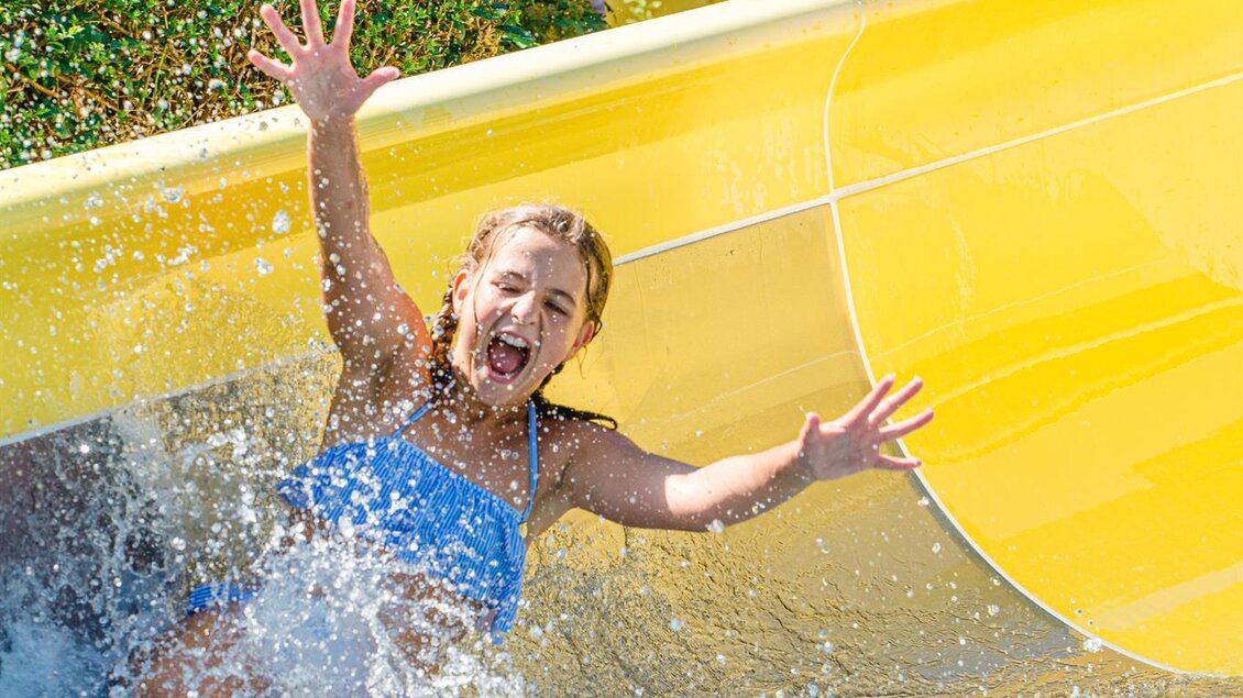 Ein Mädchen rutscht fröhlich von einer gelben Wasserrutsche ins Wasser. Spritzer und Freude sind deutlich sichtbar. | © Mias Photoart