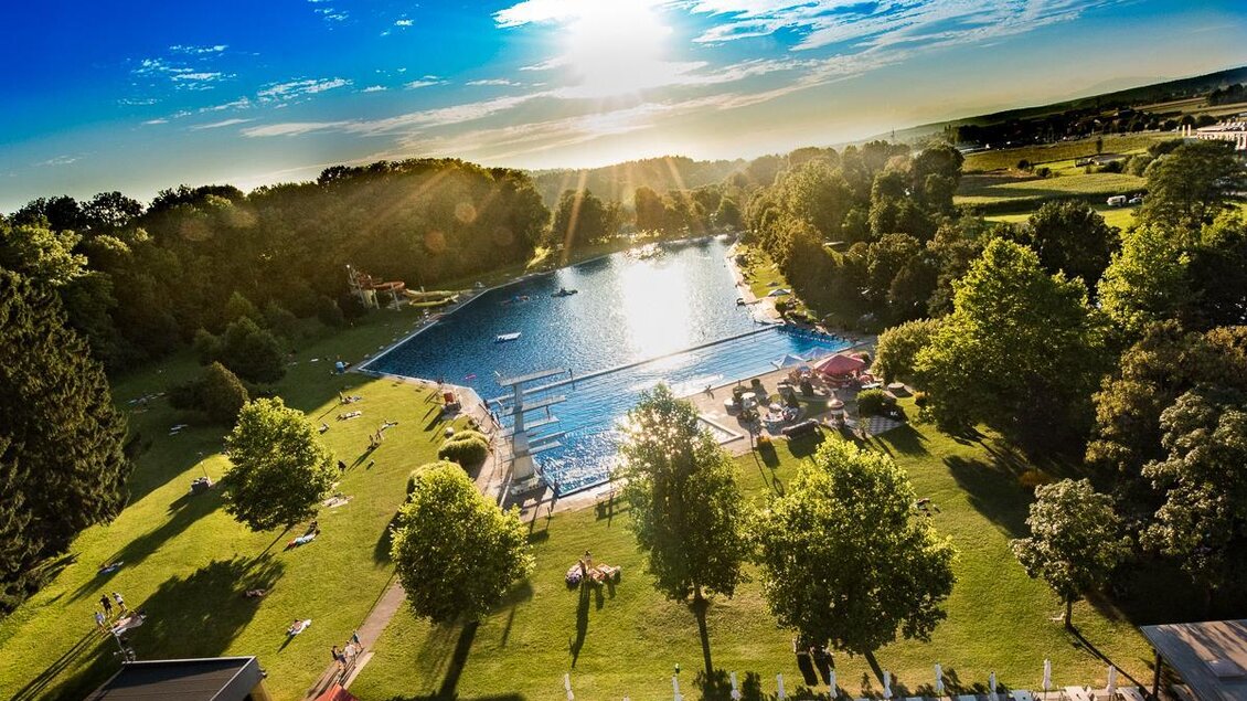 Ein schöner See umgeben von grünem Gras und Bäumen. Die Sonne scheint hell am Abendhimmel und es sind Menschen am Ufer. | © Tourismusverband Thermen- & Vulkanland