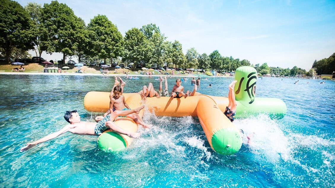 Kinder und Erwachsene haben Spaß auf einer großen schwimmenden Plattform im klaren Wasser. Die Sonne scheint und es ist ein perfekter Tag zum Spielen im Freien. | © Tourismusverband Thermen- & Vulkanland