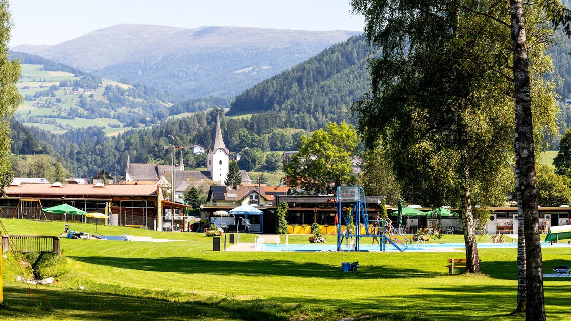 Eine malerische Landschaft mit einem grünen Rasen und einem Schwimmbereich. Im Hintergrund sind sanfte Hügel und eine Kirche zu sehen. | © TV Murau