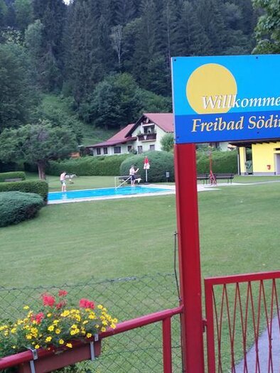 A public swimming pool with a green lawn and a pool in the background. The entrance is marked with a welcome sign. | © Johann Hiden
