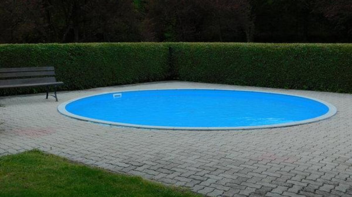 Ein runder Pool mit klarem blauem Wasser, umgeben von grünen Hecken. Neben dem Pool steht eine Holzbank auf dem gepflasterten Boden. | © www.meinbezirk.at