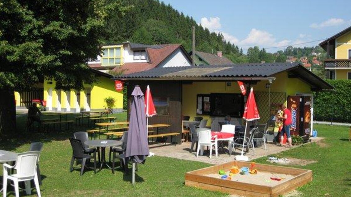 Ein kleines Café mit Außenbereich in einer grünen Umgebung. Es gibt Tische, Stühle und einen Sandkasten für Kinder. | © www.meinbezirk.at