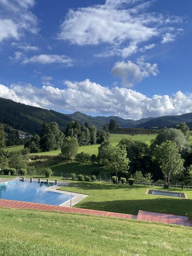 A tranquil landscape with a pool and a view of the mountains. The sky is partially cloudy, and the nature is green. | © Tourismusverband Murau