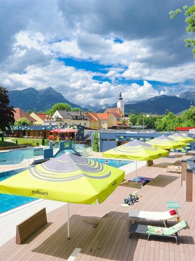 A beautiful outdoor pool with loungers and colorful sun umbrellas. In the background, mountains and a picturesque cityscape can be seen. | © TV ERZBERG LEOBEN
