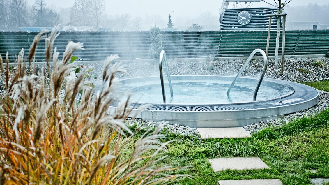 Ein Wellnessbereich mit einem warmen Whirlpool in einer natürlichen Umgebung. Dampf steigt auf, umgeben von Gräsern und einer ruhigen Gartenlandschaft. | © Holding Graz - Lupi Spuma