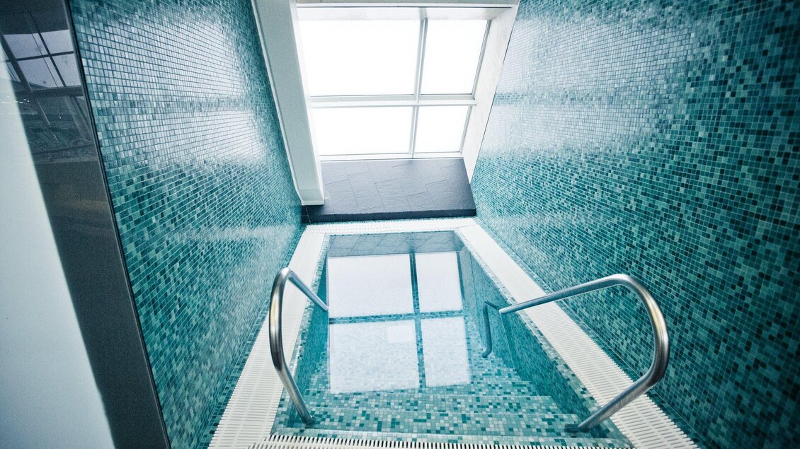 Ein modernes Schwimmbecken mit blauen Fliesen und einem großen Fenster. Die glänzende Wasseroberfläche spiegelt den Raum wider. | © Holding Graz - Lupi Spuma