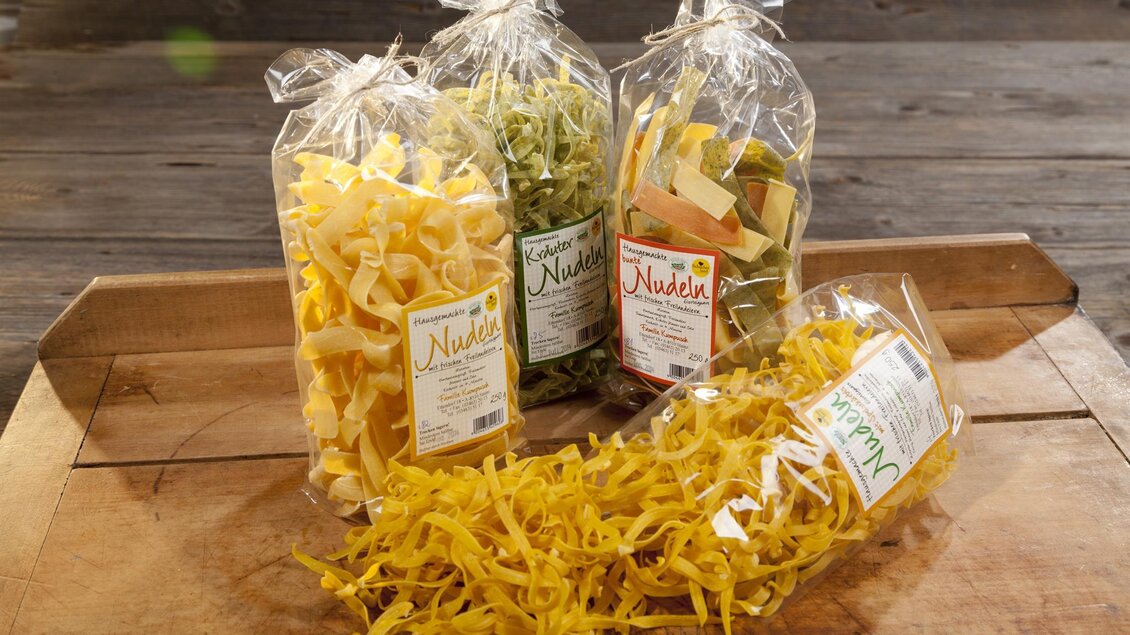Vier verschiedene Sorten von Nudeln in transparenter Verpackung auf einem Holztisch. Die Farben reichen von gelb bis grün, ideal für eine leckere Pasta-Mahlzeit. | © Schilcherland Spezialitäten