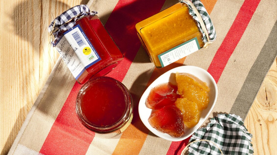 Drei Gläser Marmelade stehen auf einem gestreiften Tischläufer. Daneben liegt eine Schale mit verschiedenen Fruchtgummis. | © Schilcherland Spezialitäten