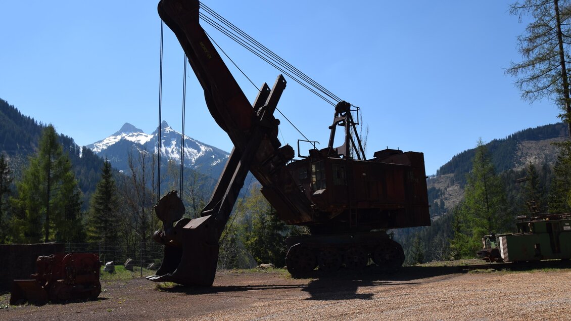 Eine große Baggermaschine steht im Vordergrund, umgeben von Bergen und Bäumen. Der Himmel ist klar und blau. | © Abenteuer Erzberg