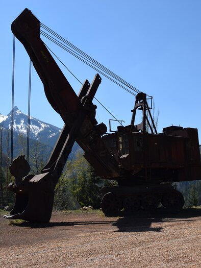 A large excavator is in the foreground, surrounded by mountains and trees. The sky is clear and blue. | © Abenteuer Erzberg