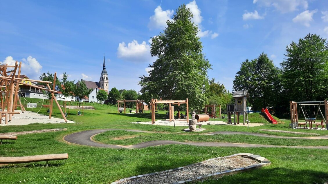 Ein schöner Spielplatz mit verschiedenen Spielgeräten in einer grünen Umgebung. Im Hintergrund ist eine Kirche mit einem markanten Turm zu sehen. | © Marktgemeinde Pischelsdorf