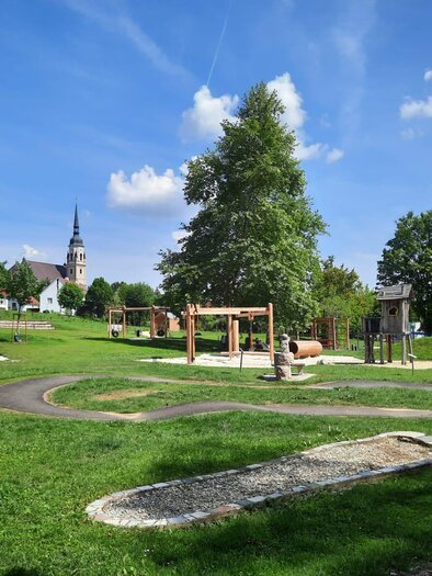 Ein schöner Spielplatz mit verschiedenen Spielgeräten in einer grünen Umgebung. Im Hintergrund ist eine Kirche mit einem markanten Turm zu sehen. | © Marktgemeinde Pischelsdorf