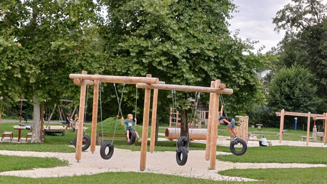 Ein Spielplatz mit Holzschaukeln und einer Rutsche. Grüne Wiesen und Bäume umgeben den Bereich. | © Marktgemeinde Pischelsdorf