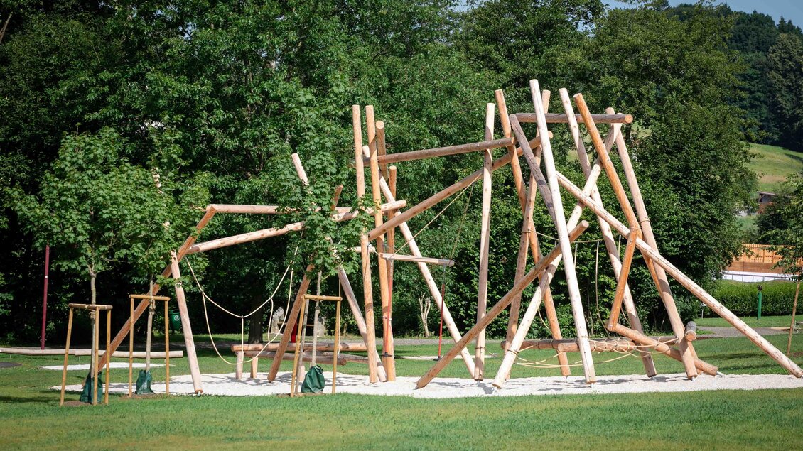 Ein Spielplatz aus Holz mit verschiedenen Klettermöglichkeiten und Schaukeln. Umgeben von grünen Bäumen und einer Wiese. | © fotografiebrunner.com