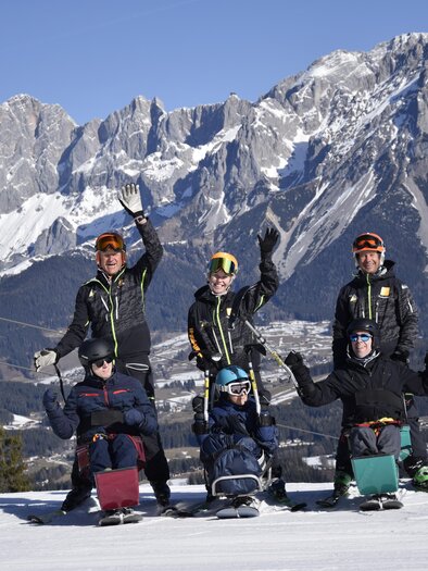 A group of skiers is sitting on sleds in the snow, surrounded by impressive mountains. Everyone is smiling and waving at the camera. | © Lisa-Marie Reiter