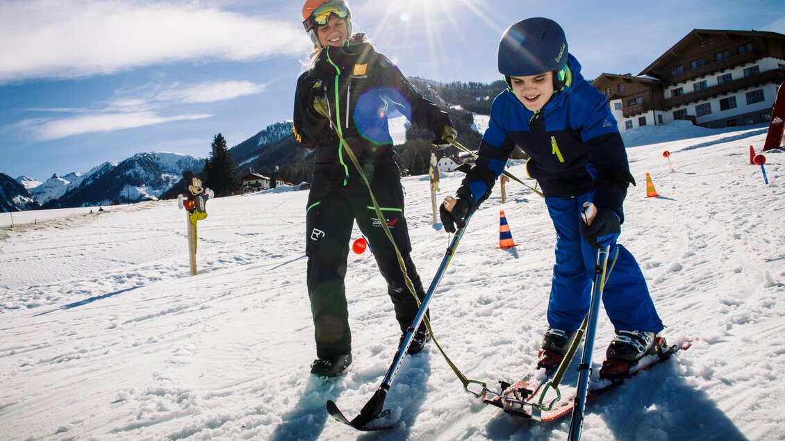 Ein Skilehrer hilft einem kleinen Jungen beim Skifahren auf einer schneebedeckten Piste. Die Sonne scheint und im Hintergrund sind Berge und Gebäude sichtbar. | © Lisa Marie Reiter