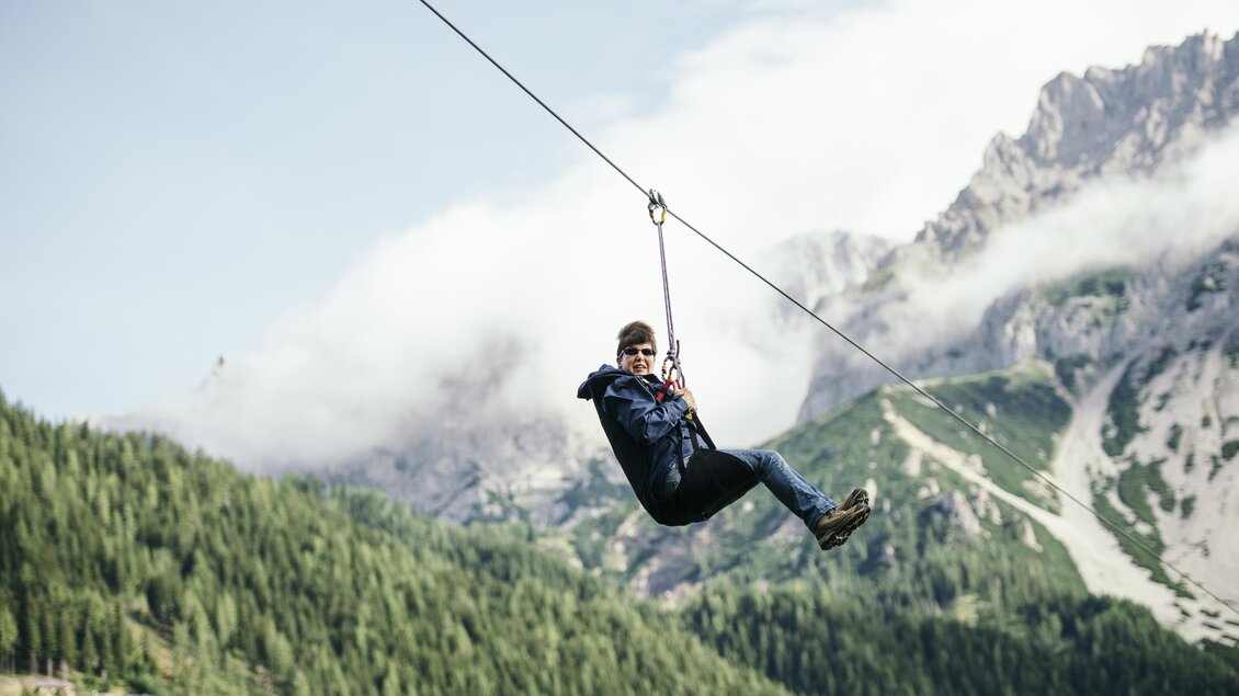 Eine Person fährt an einer Seilrutsche durch eine grüne Berglandschaft. Im Hintergrund sind Wolken und hohe Berge zu sehen. | © Lisa Marie Reiter