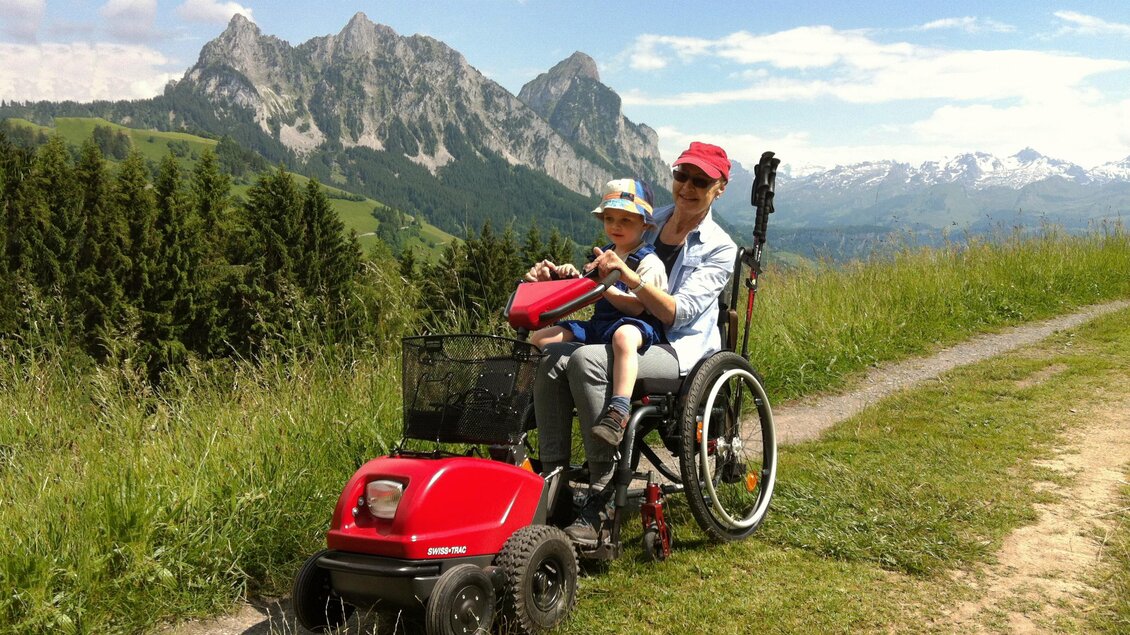 Ein Kind sitzt auf einem roten Freizeitbuggy, der von einem Erwachsenen im Rollstuhl geschoben wird. Im Hintergrund sind malerische Berge und ein blauer Himmel zu sehen. | © Freizeit-PSO