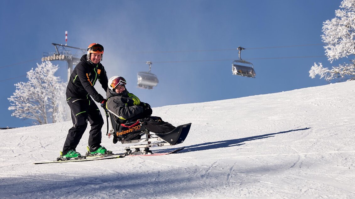 Ein Skifahrer zieht einen Mann in einem Schlitten über eine schneebedeckte Piste. Im Hintergrund sind Skilifte und verschneite Bäume zu sehen. | © Lisa-Marie Reiter