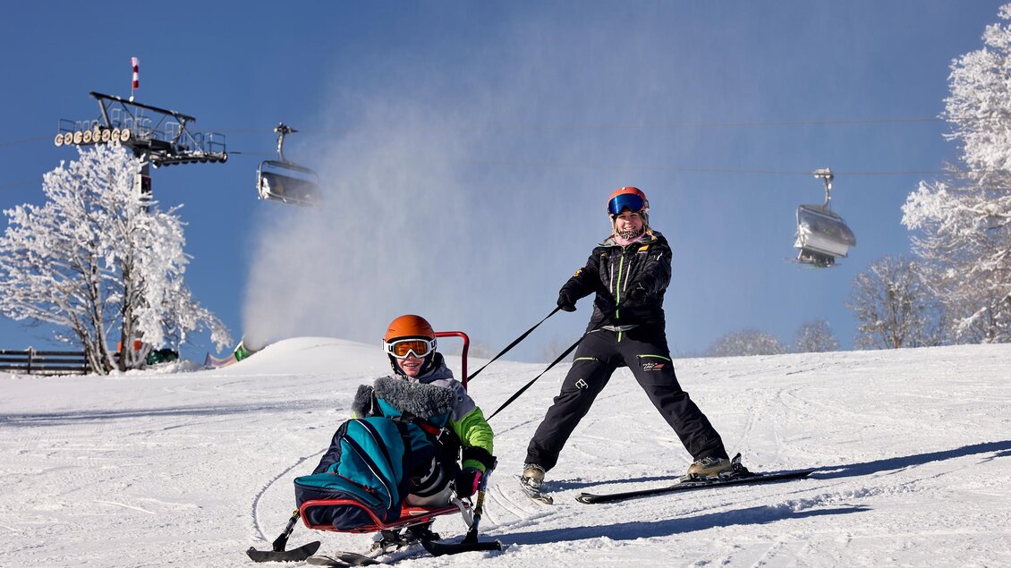 Zwei Kinder auf einer Skipiste im Schnee. Eines zieht das andere auf einem Schlitten, umgeben von schneebedeckten Bäumen und einem blauen Himmel. | © Lisa-Marie Reiter