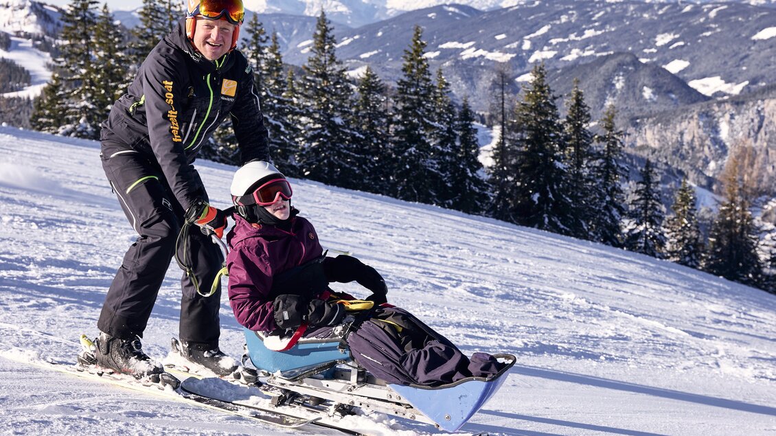 Ein Skifahrer zieht einen Schlitten mit einem passierenden Beifahrer den Hang hinunter. Im Hintergrund sind schneebedeckte Berge und Nadelbäume zu sehen. | © Lisa-Marie Reiter