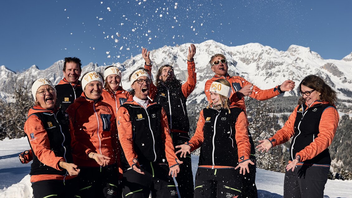 Eine Gruppe von fröhlichen Skifahrern steht im Schnee und wirft mit Schnee um sich. Im Hintergrund sind schneebedeckte Berge zu sehen. | © Lisa-Marie Reiter