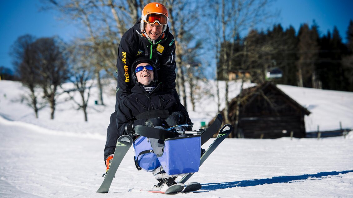 Eine Person fährt in einem Schlitten, während eine andere Person hinter ihnen steht und sie unterstützt. Es ist ein sonniger Tag im Schnee mit Bäumen im Hintergrund. | © Lisa-Marie Reiter