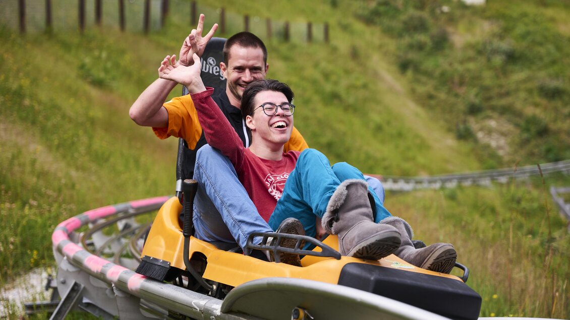 Zwei Personen sitzen auf einer Rodelbahn und genießen den Fahrspaß. Beide lachen und zeigen mit den Händen ein Zeichen des Glücks. | © Lisa-Marie Reiter