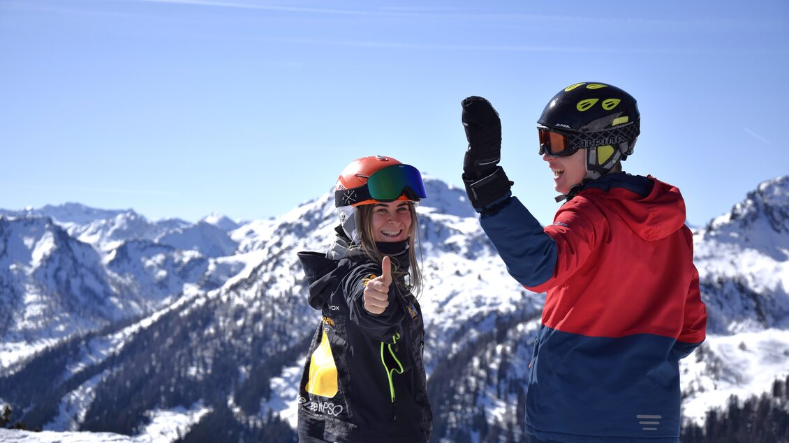Zwei Skifahrer stehen auf einem schneebedeckten Berggipfel und freuen sich. Im Hintergrund sind beeindruckende Berglandschaften und ein klarer blauer Himmel zu sehen. | © Lisa-Marie Reiter