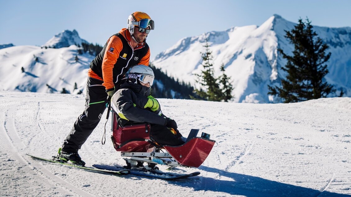 Ein Skifahrer hilft einer Person im Schlitten auf einer verschneiten Piste. Im Hintergrund sind majestätische Berge und ein klarer blauer Himmel zu sehen. | © Lisa Marie Reiter
