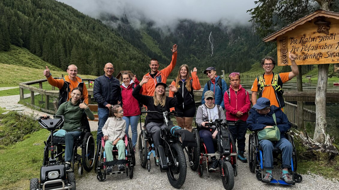 Eine Gruppe von Menschen in Rollstühlen steht fröhlich in einer malerischen Landschaft mit Bergen im Hintergrund. Sie posieren vor einem Holzschild und genießen die Natur. | © Lisa Marie Reiter