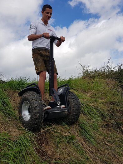 A young man is standing on an all-terrain scooter on a hill. In the background, there is grass and a blue sky with clouds. | © Hotel-Restaurant Perschler