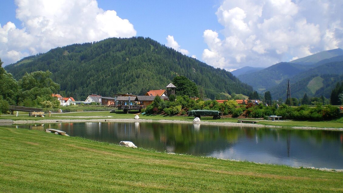 Eine ruhige Landschaft mit einem See und grünen Wiesen. Im Hintergrund sind sanfte Hügel und eine malerische Dorfansicht sichtbar. | © Anita Fössl