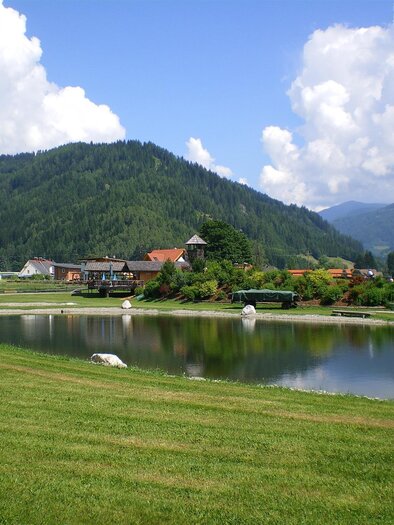 A tranquil landscape with a lake and green meadows. In the background, gentle hills and a picturesque village view are visible. | © Anita Fössl