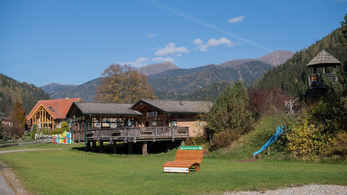 Eine malerische Landschaft mit einem Holzhaus und sanften Bergen im Hintergrund. Im Vordergrund gibt es eine weitläufige Grünfläche und Spielgeräte. | © Anita Fössl