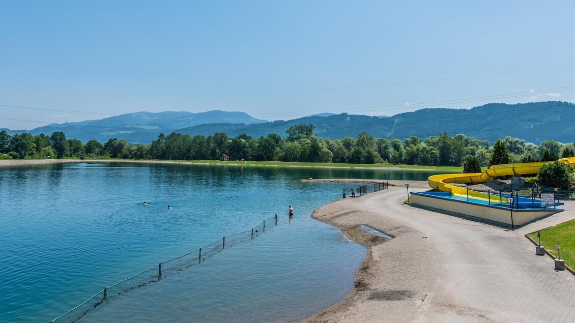 Ein klarer See mit einem Sandstrand und einem großen Wasserrutschenbereich. Im Hintergrund sind grüne Bäume und Berge zu sehen. | © Freizeitanlage Zechner