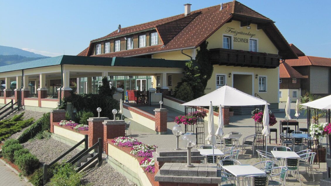 Ein Restaurant mit einer einladenden Terrasse und Tischen im Freien. Im Hintergrund ist ein charmantes Gebäude mit einem roten Dach zu sehen. | © Freizeitanlage Zechner