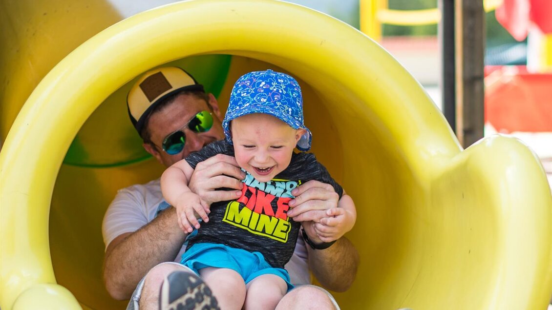 Ein Erwachsener und ein kleines Kind sitzen gemeinsam in einer Rutsche. Beide lachen und haben Spaß beim Spielen im Freien. | © Freizeitanlage Zechner