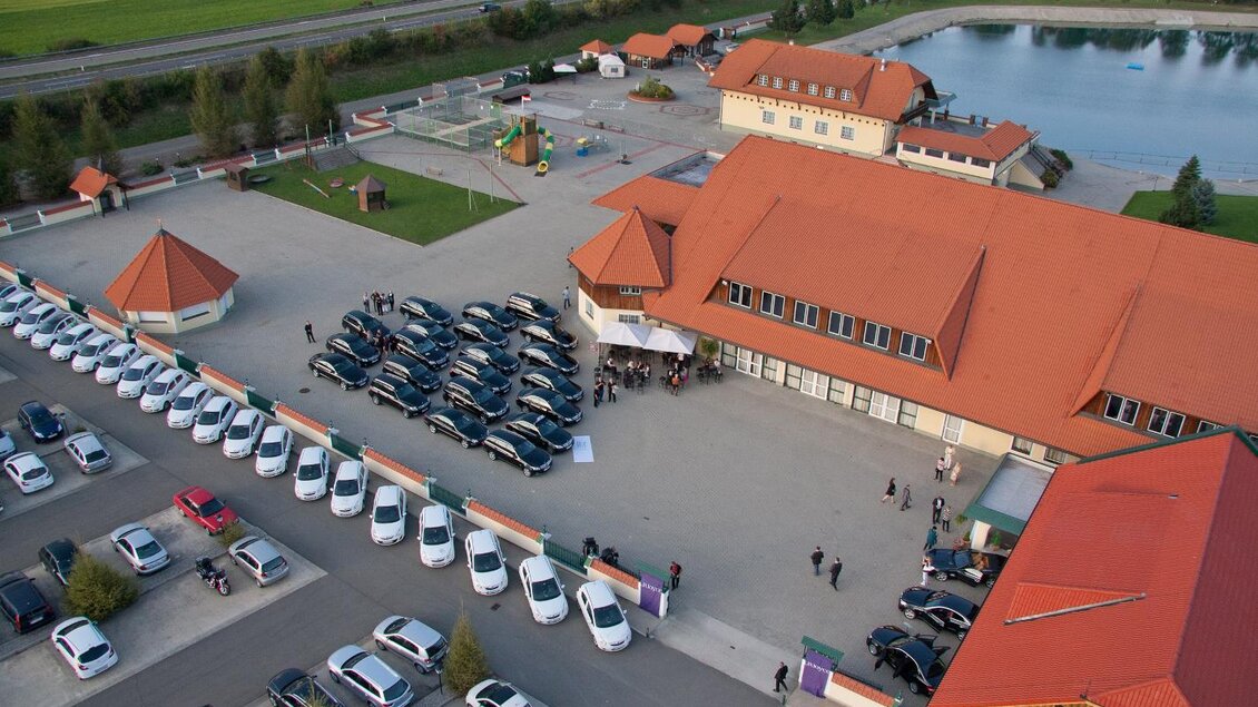 Eine Luftaufnahme eines Parkplatzes mit vielen Autos und einem Gebäude mit orangefarbenem Dach. Im Hintergrund ist ein Teich und grüne Landschaft zu sehen. | © Freizeitanlage Zechner